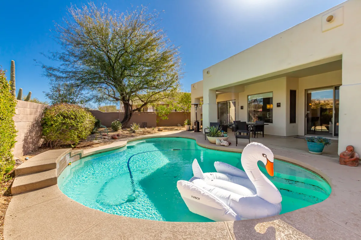 A vibrant backyard scene featuring a swimming pool with an inflatable swan, surrounded by lush landscaping. The pool area is clearly designed for family fun, with plenty of open space for children to swim and play under the sun.
