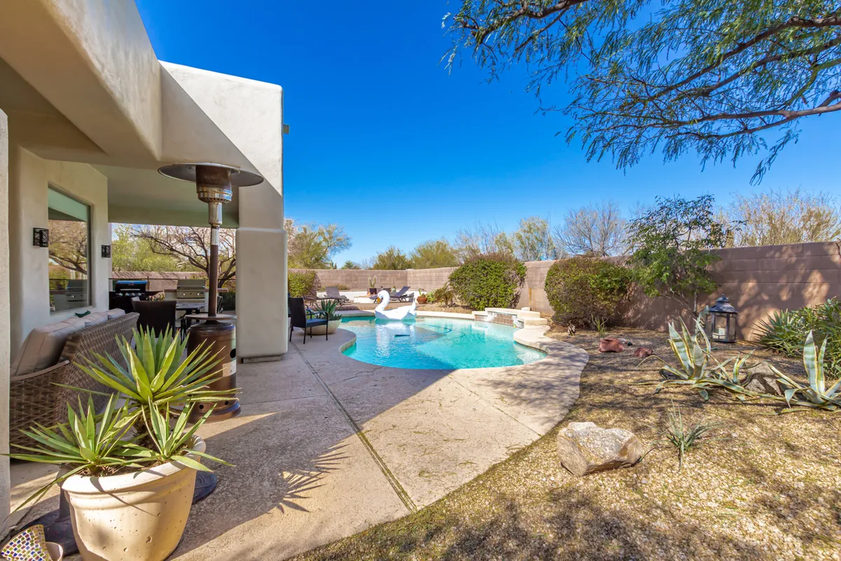 A tranquil backyard oasis featuring a sparkling swimming pool surrounded by lounge chairs, an inviting outdoor seating area, and lush potted plants. The scene is bathed in warm Arizona sunlight, ideal for relaxation or socializing, with the pool as the focal point and hints of the private, exclusive setting.