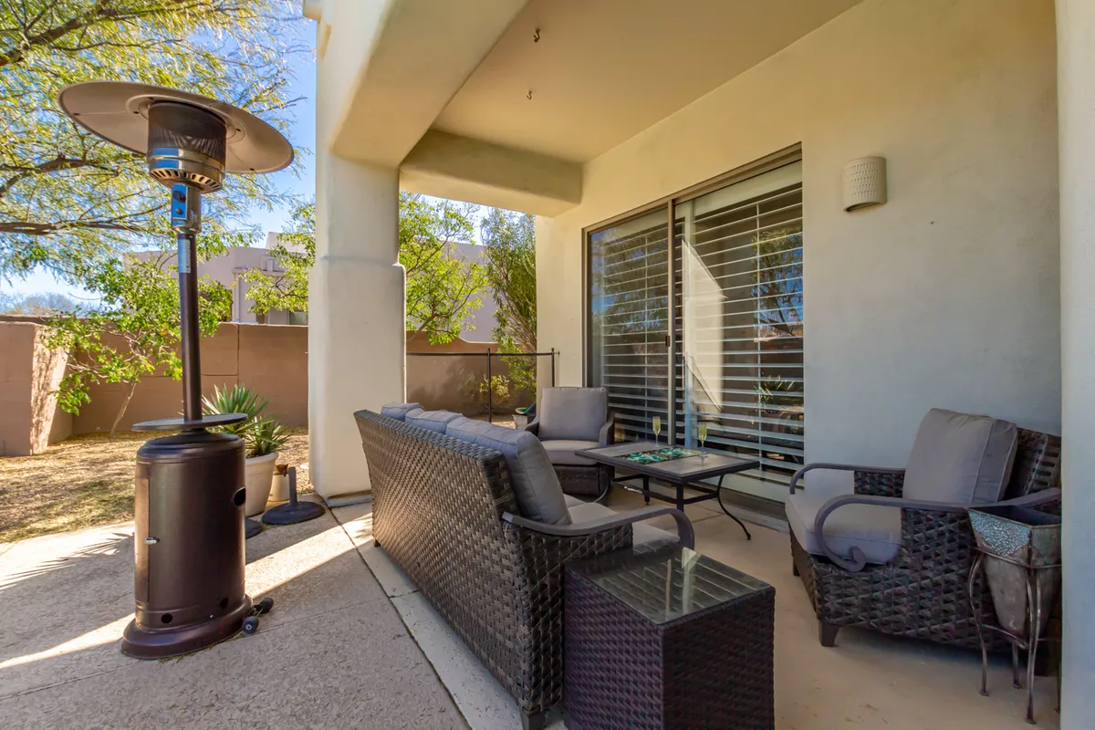 A cozy outdoor seating area with wicker furniture, an outdoor heater, a glass table, and potted plants. The setting is intimate, surrounded by desert landscaping, inviting relaxation and socialization. Soft morning or evening light enhances the tranquil atmosphere, perfect for unwinding or enjoying a cup of coffee while taking in the desert views.