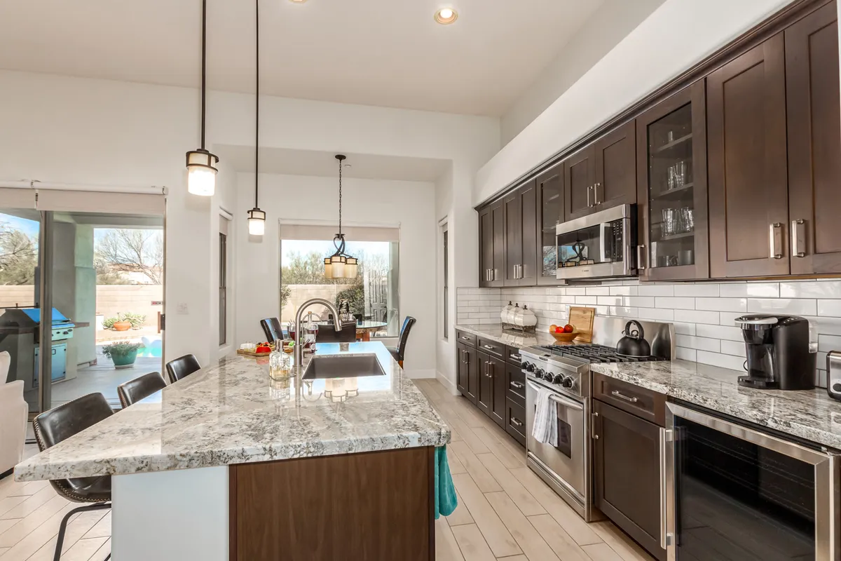 The image displays a kitchen with sleek granite countertops, dark wood cabinetry, and modern stainless appliances. In the background, a dining area bathed in natural light is visible, offering a seamless transition between cooking and dining spaces.