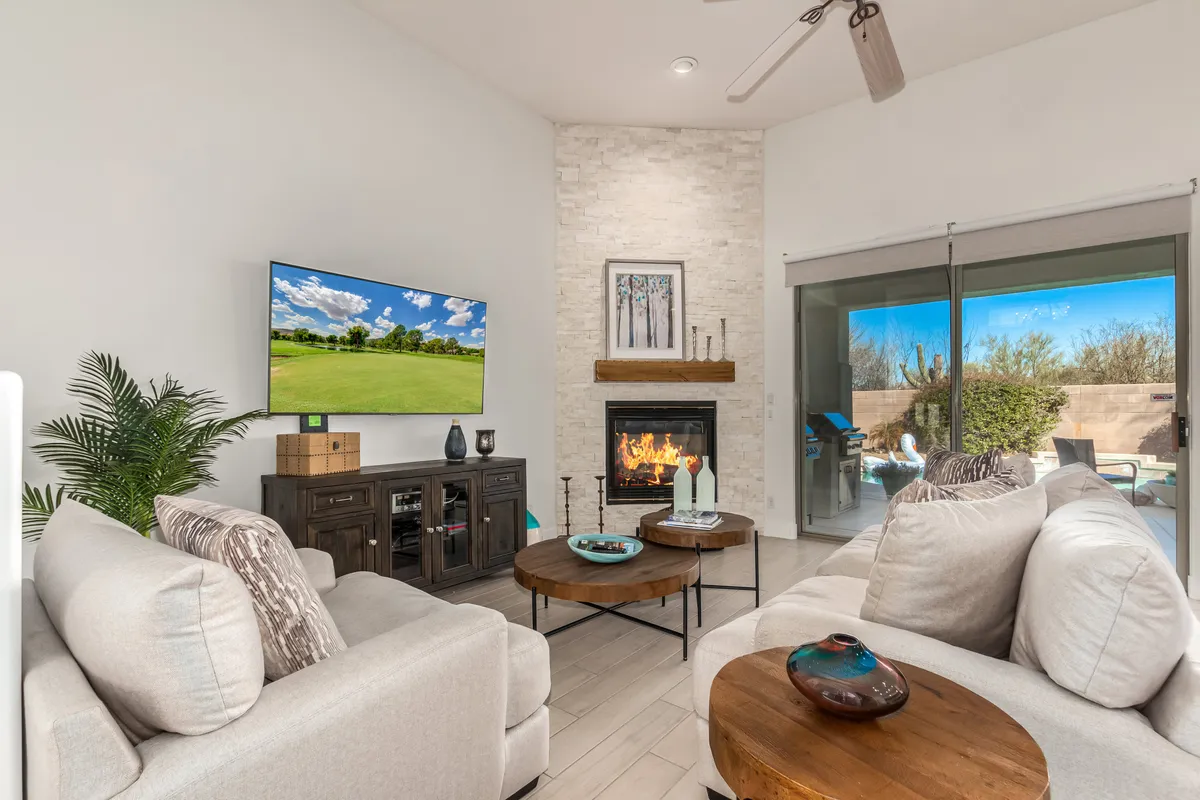 A welcoming living room with comfortable sofas arranged around a modern fireplace and a large television. A sliding glass door fills the space with natural light and offers a glimpse of the desert landscape outside. Coffee tables provide a casual gathering spot, and the decor feels warm and inviting.