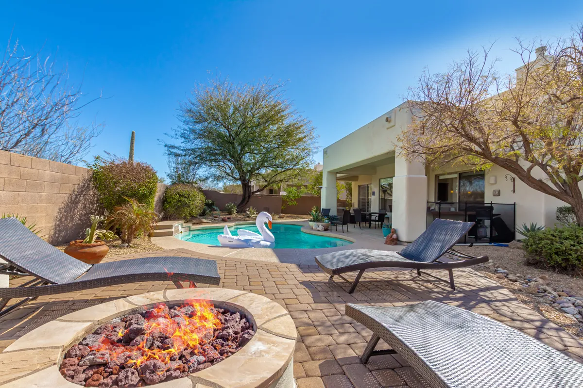The image showcases a stunning backyard oasis at sunset: a glimmering swimming pool bordered by plush lounge chairs, whimsical swan floaties drifting on the water, and a glowing fire pit surrounded by inviting seating. Lush desert landscaping frames the space, creating an intimate yet spacious area ideal for relaxation or gathering with friends and family.