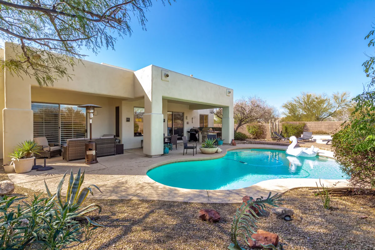 This exterior image showcases a sparkling swimming pool surrounded by desert landscaping and an inviting outdoor seating area. The grill area is visible in the background, and desert plants accentuate the tranquil oasis feel. The setting appears perfect for swimming, sunbathing, and alfresco dining, all within a private, gated atmosphere.