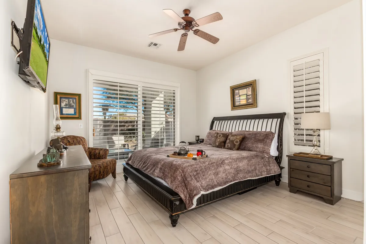A serene bedroom with plush bedding, wooden furniture, a window with shutters allowing in natural light, and a television mounted on the wall. The ambiance is calm and inviting for relaxation or restful sleep.