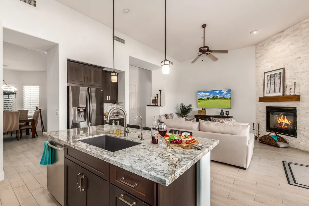 A spacious, cozy area where the kitchen, living room, and dining space come together. A comfy sofa faces a fireplace and a TV, while a large island and dining area are visible in the same view. Natural light and tasteful decor make the space feel inviting and warm.